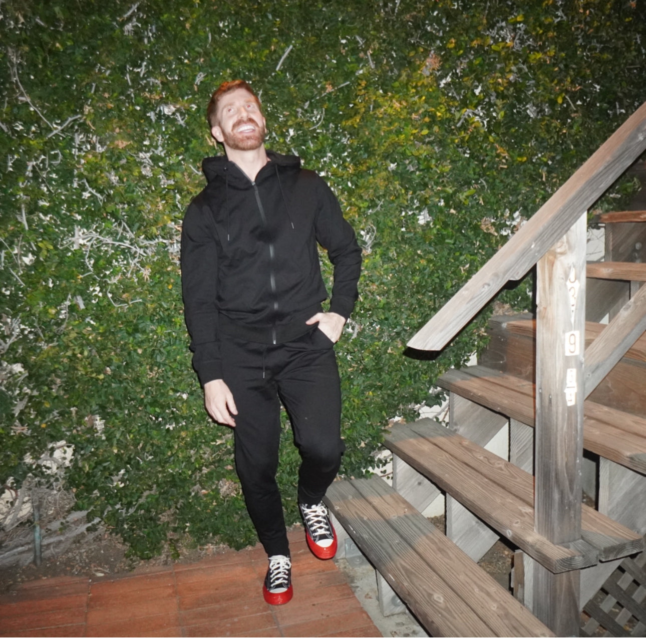 A man in black sweatshirt and sneakers laughing standing by stairs.